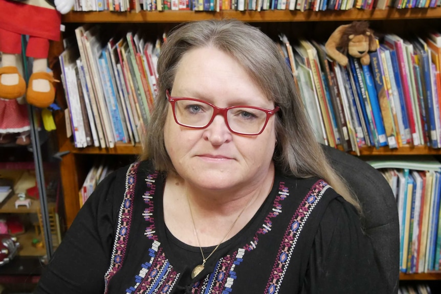 A woman with red glasses sits in front of a bookshelf.