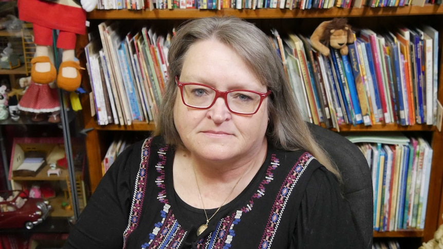 A woman with red glasses sits in front of a bookshelf.