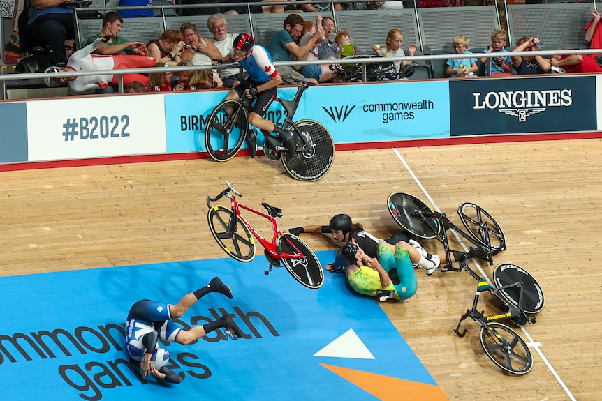 Three riders lay on the ground, one collects himself and one lays horizontally in the crowd after a crash