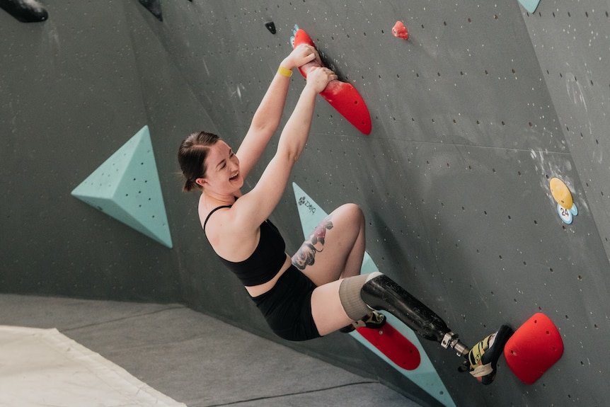 Sarah Larcombe hangs on a climbing wall with red bars.