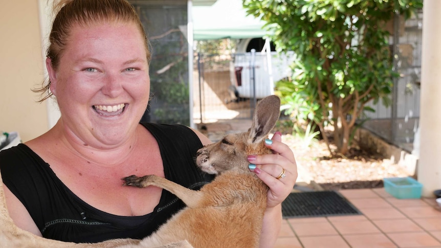 A woman in a black singlet smiles at the camera holding a young kangaroo.