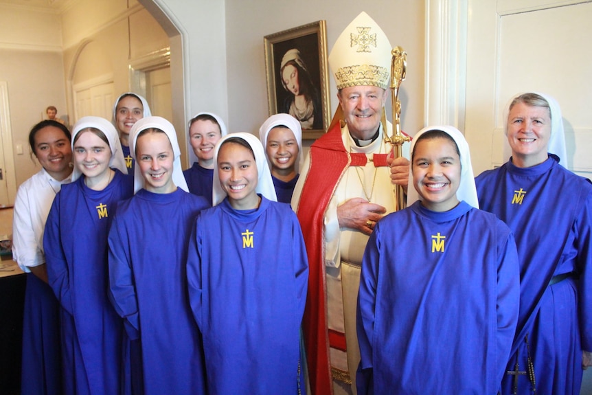 An archbishop wearing robes and hat, holding a staff, standing with a group of nuns wearing blue habits and white veils