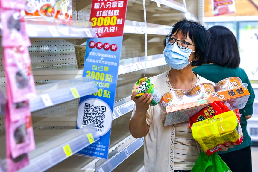 A woman in a face mask with her arms filled with food scans an empty supermarket shelf