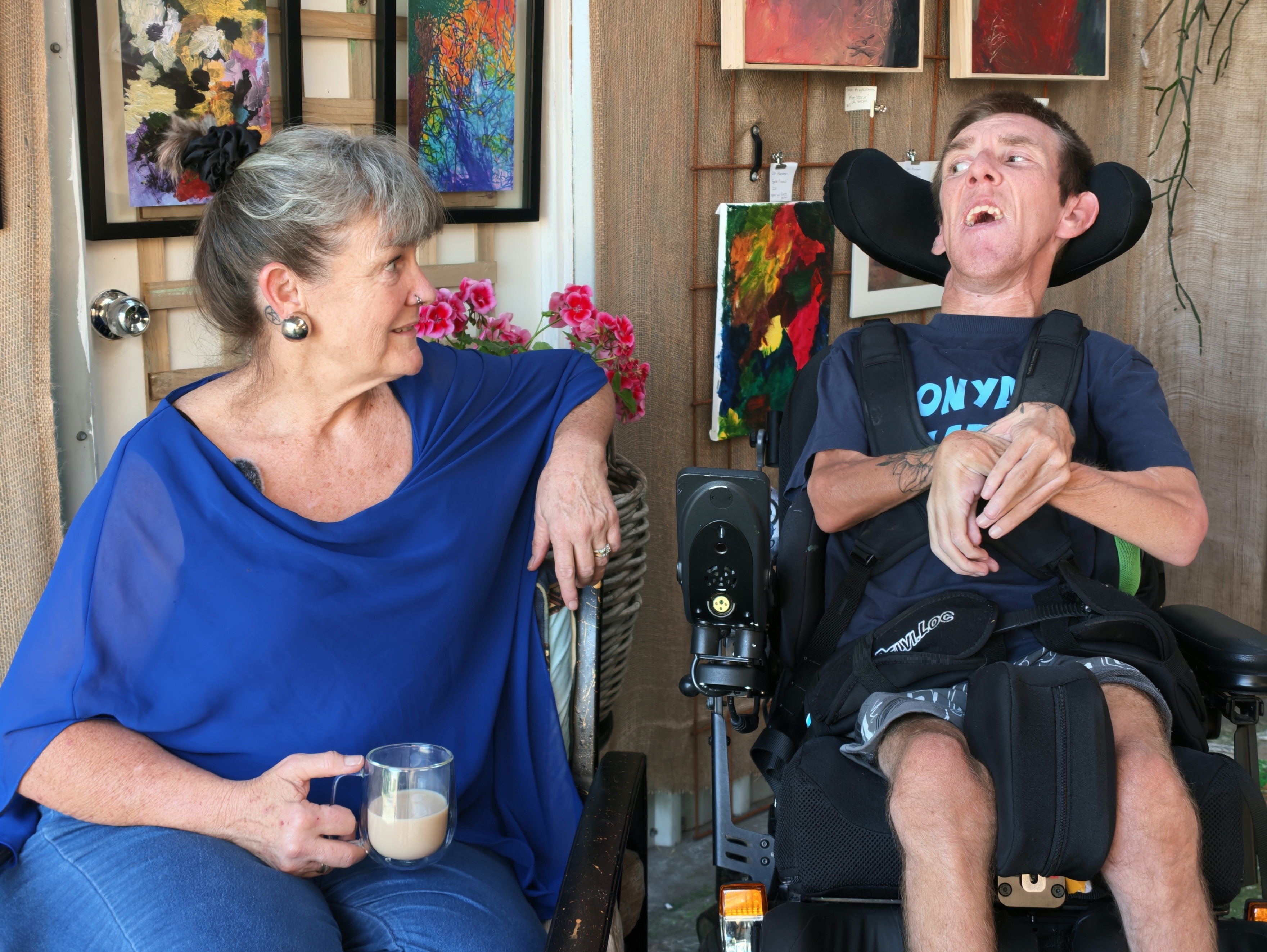 Wendy and Colin Hanssen sit side by side in front of artwork on a wall.