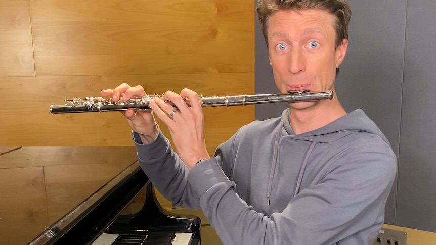 Comedian Sammy J plays flute while sitting at a grand piano. His eyes are open very wide.