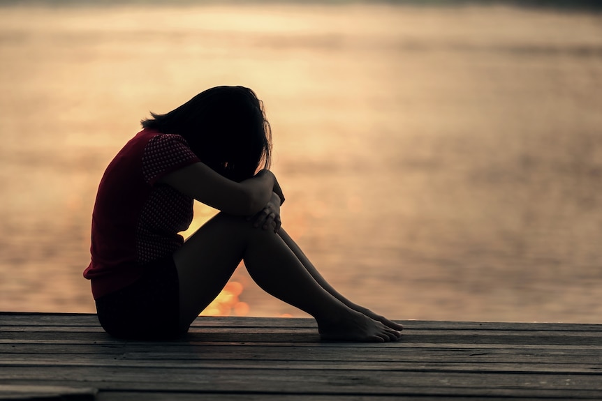 Silhouette of a slim woman sitting on a wooden dock or jetty next to water in low light, with her head in her hands.