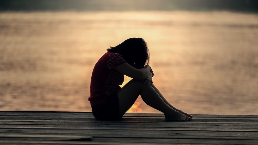 Silhouette of a slim woman sitting on a wooden dock or jetty next to water in low light, with her head in her hands.