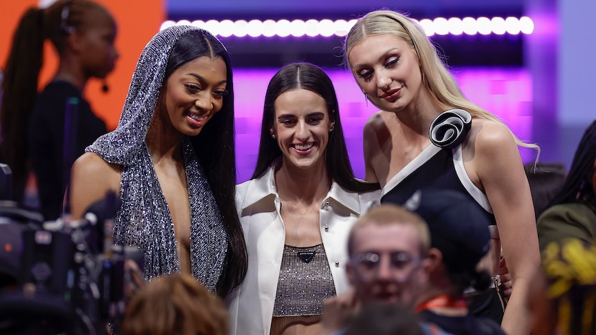 Three women smile for a camera while wearing designer clothing. 