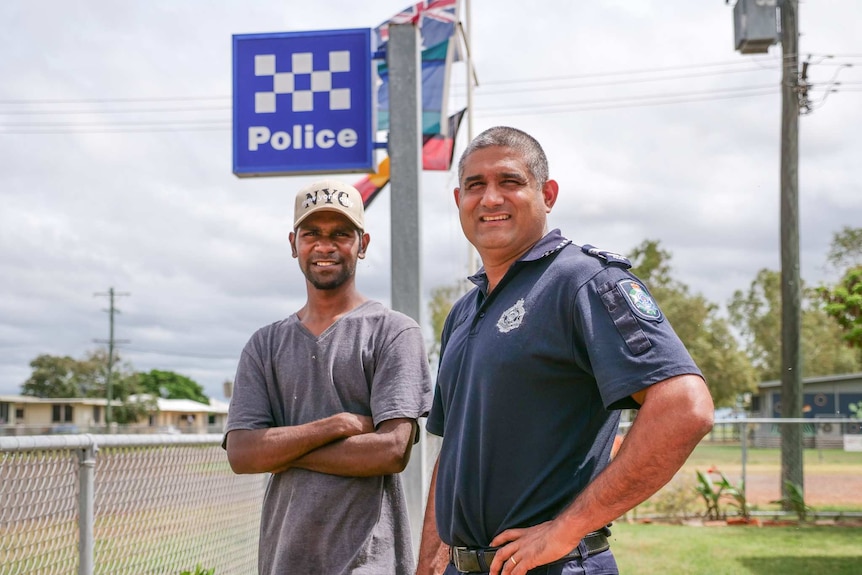 A young indigenous man stands, arms crossed, next to a police officer. Australian and Indigenous flags are in the background.