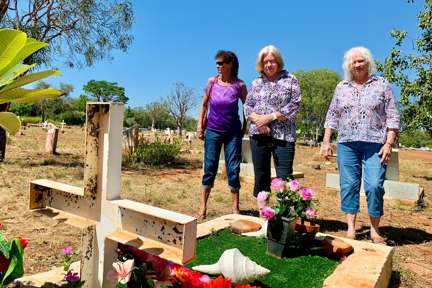 Three women stand looking at a grave at a sunny bush cemetery