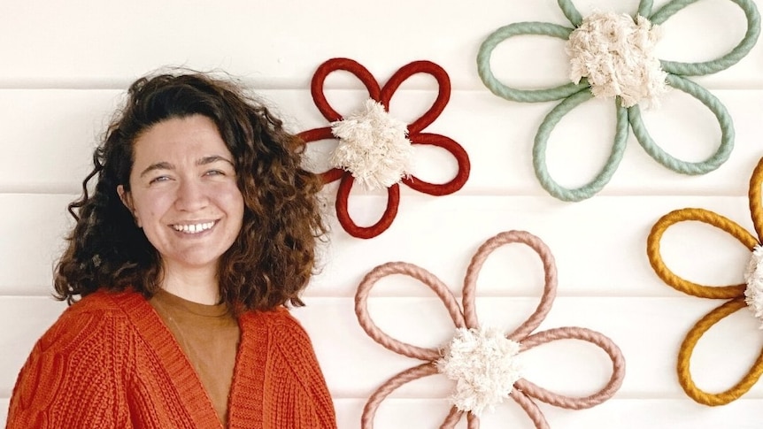 A woman in a burnt-orange cardigan stands next to a display of flower-shaped macrame wall hangings.