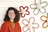 A woman in a burnt-orange cardigan stands next to a display of flower-shaped macrame wall hangings.