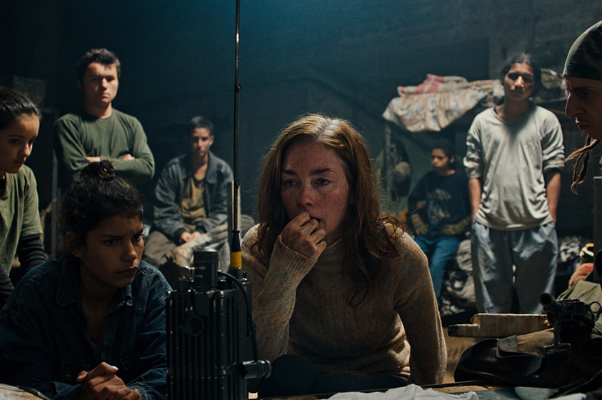 A woman with red hair ponders in front of radio in bunker surrounded by 5 teen boys and 2 teen girls looking serious.