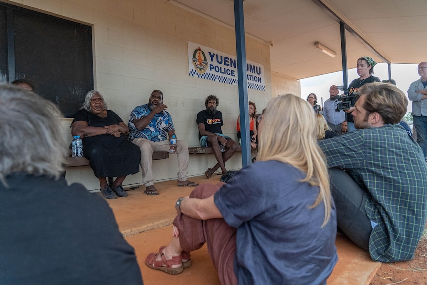 A group of people sit on the ground outside the Yuendumu police station. Faded red handprints can be seen on the wall.