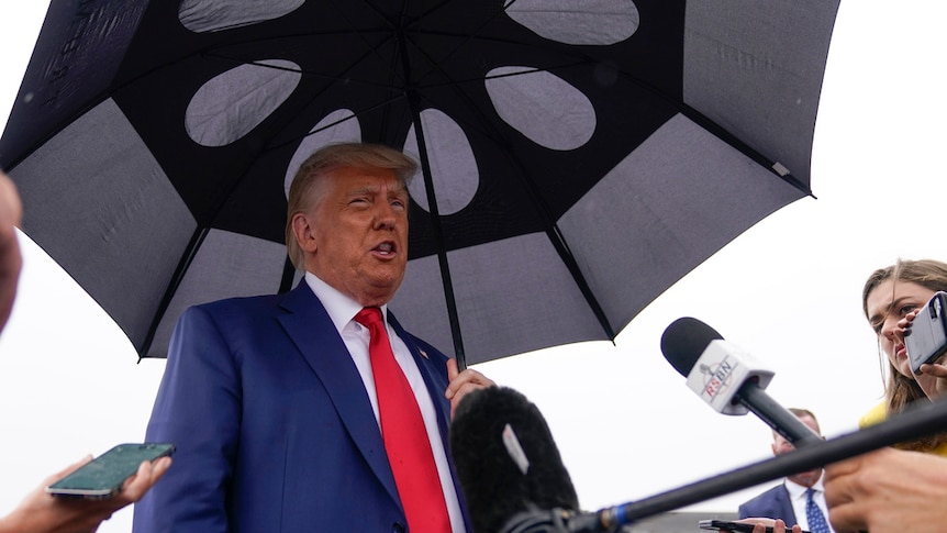 Donald Trump, holding an umbrella in the rain, speaks to reporters at an airport.