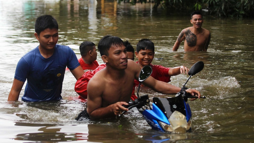 Six men and toys walk and push a motorbike through floodwater