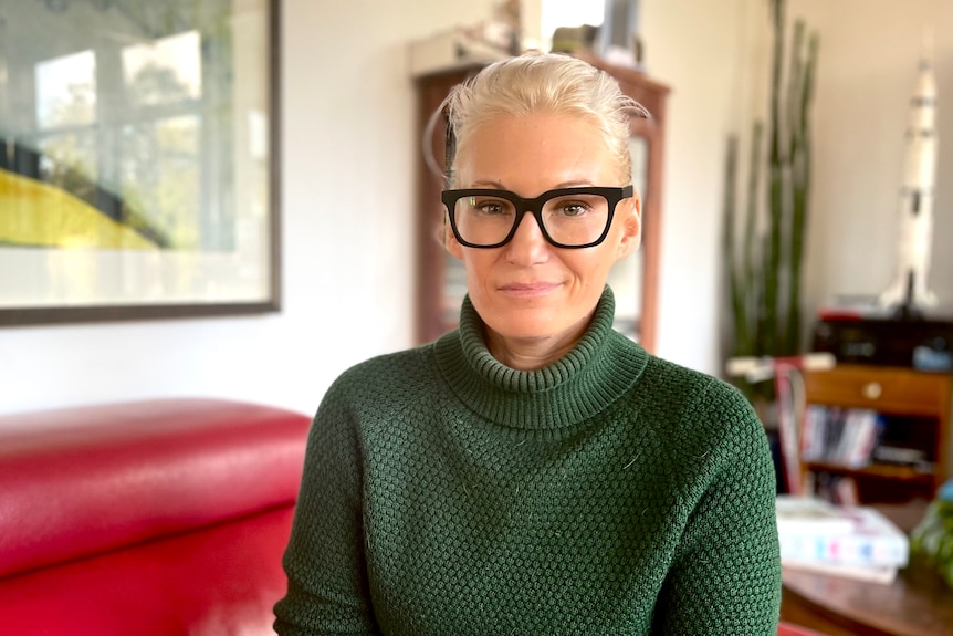 Blonde lady with hair pulled back, wearing funky black glasses and a green jumper smiles slightly at camera 