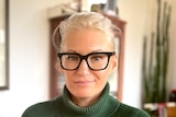 Woman with hair pulled back, wearing funky black glasses and a green jumper smiles slightly at camera 