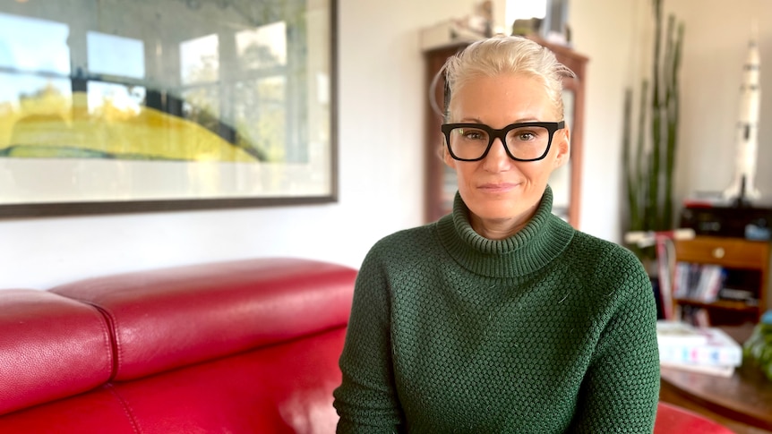 Woman with hair pulled back, wearing funky black glasses and a green jumper smiles slightly at camera 