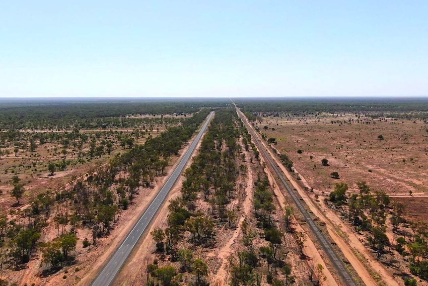 Aerial view of a highway with parallel train tracks in the outback. Red dirt and sparse trees.