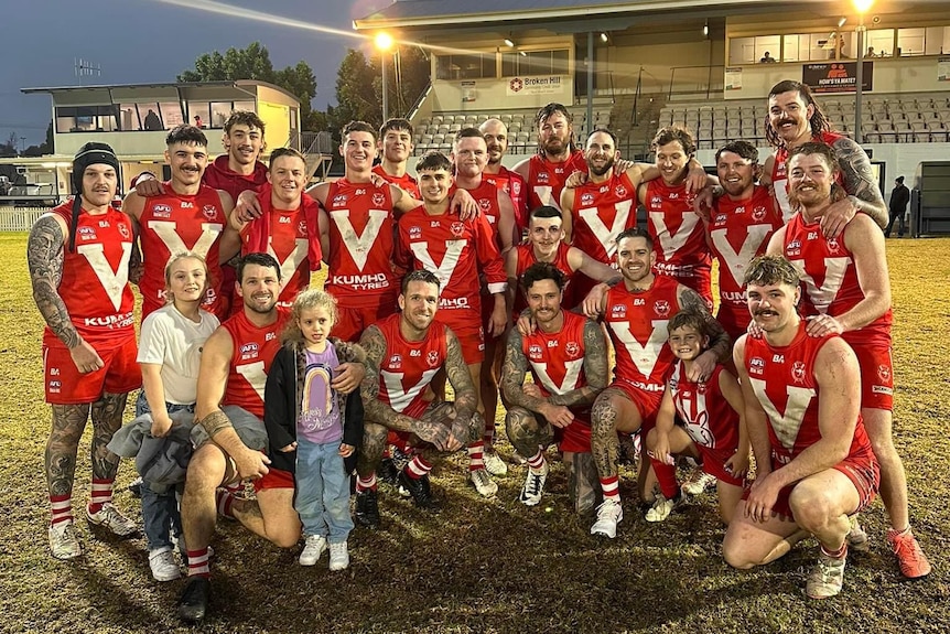 A group of men in red and white guernseys with a couple of children posing on an afl oval in front of some stands.