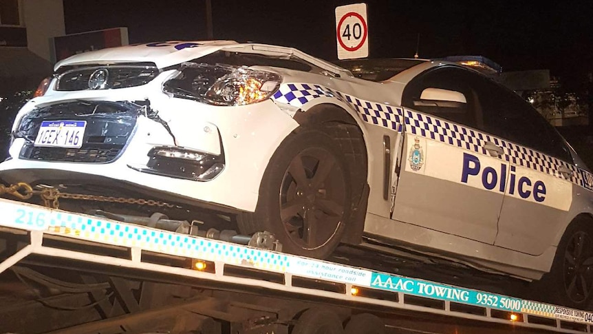 A WA Police car sits on a flatbed truck with its front smashed up at night.