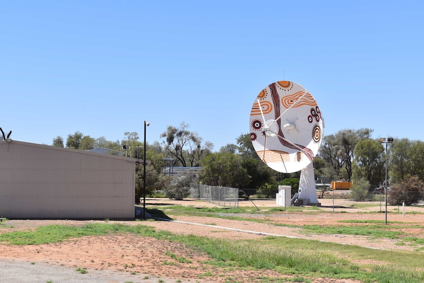 A painted satellite sits in a fenced off area next to a building.