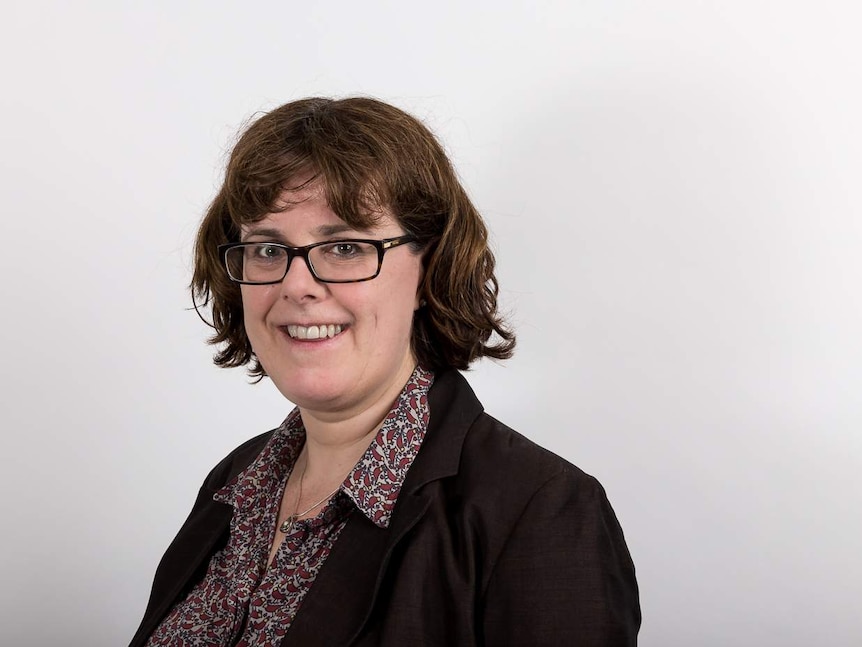 A woman with shoulder length brown hair and glasses in professional clothes smiling at a camera