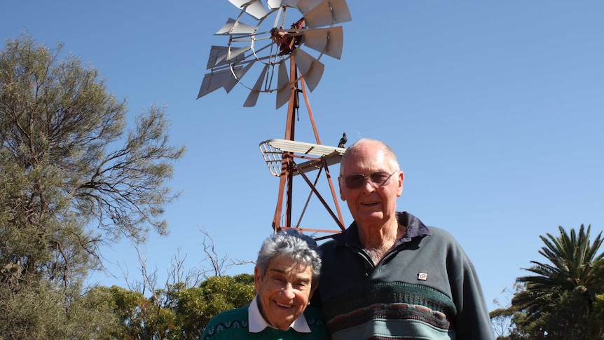 An older couple stand smiling with their arms around each other's shoulders outdoors in front of a windmill.
