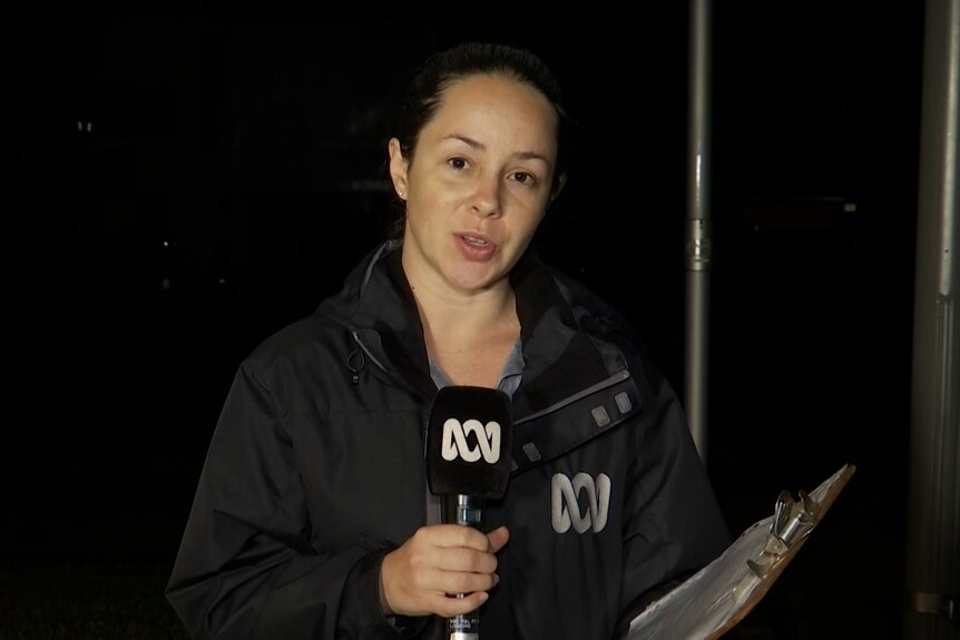 A woman standing in the dark with an ABC microphone and rain jacket.