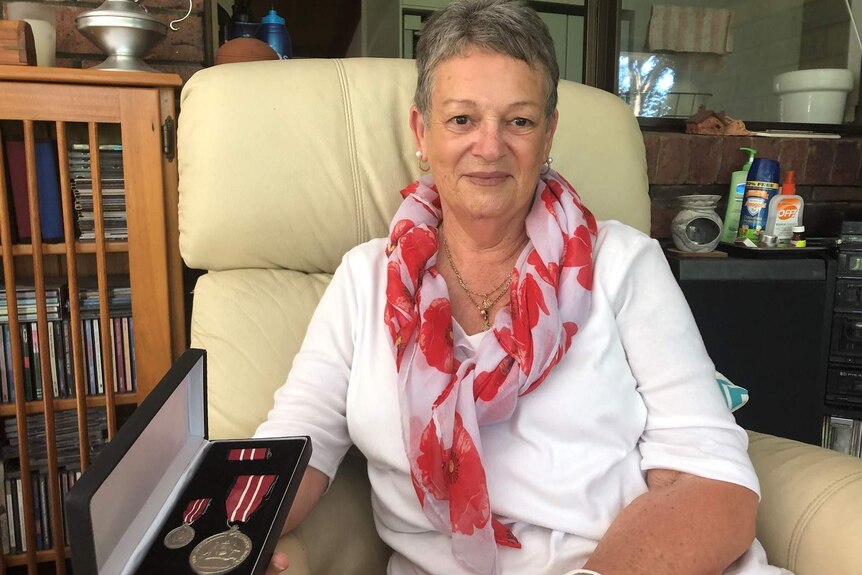 Deirdre Elliott sits in an armchair, holding her Army service medal. She is wearing a scarf with poppies on it.