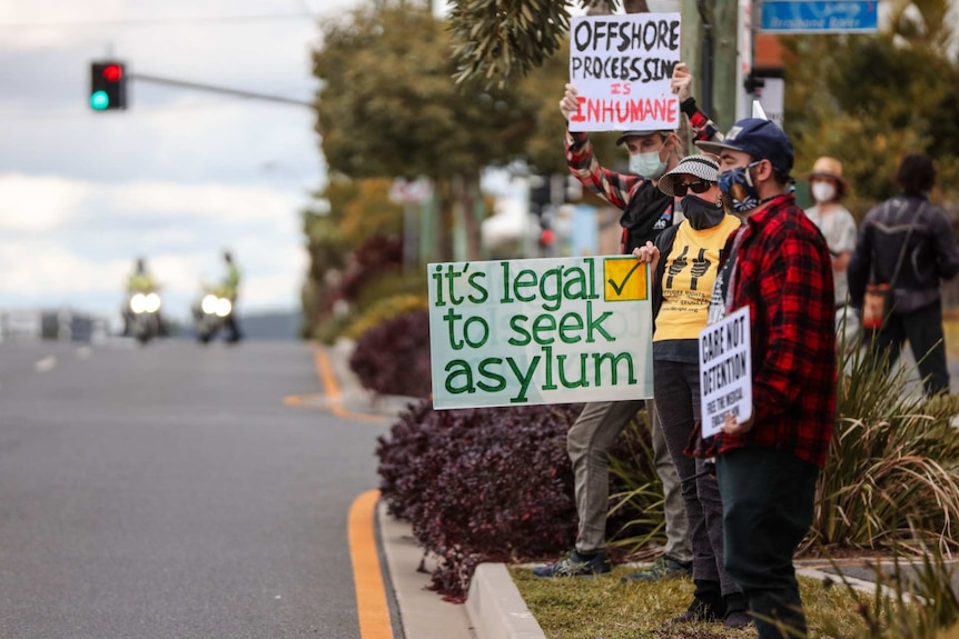 Protesters stand on the side of a street holding placards that say "it's legal to seek asylum".