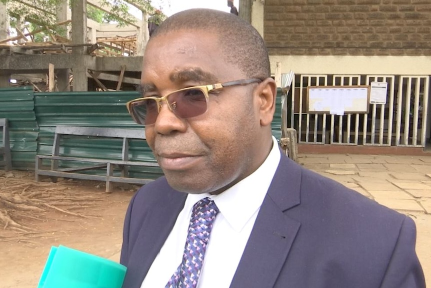 Wilfred Nderitu stands outside court looking past the camera while wearing a suit and glasses.