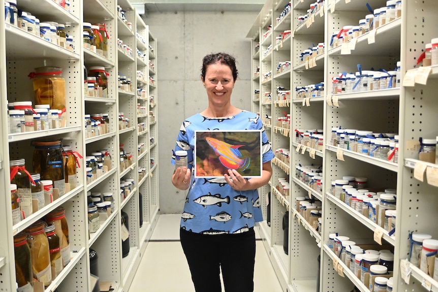 A woman standing between specimen collection shelves holding a photo of a tropical fish