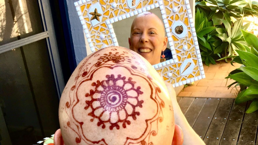 A bald woman with an intricate henna crown smiles into a mirror.