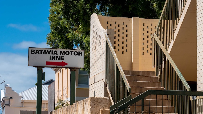 A sign says 'Batavia Motor Inne' with an arrow that points to a run down staircase to enter the motel. The sky is blue.