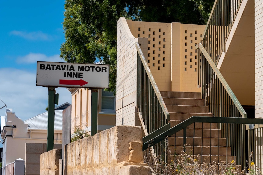 A sign says 'Batavia Motor Inne' with an arrow that points to a run down staircase to enter the motel. The sky is blue.