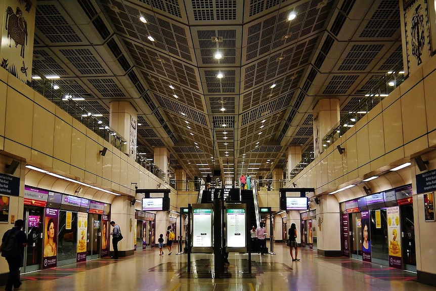 You see a shopping mall-like underground train station with lit information screens lining a vast walkway.
