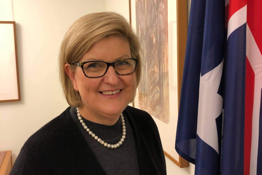 Middle aged woman with glasses stands in front of Australian flag.