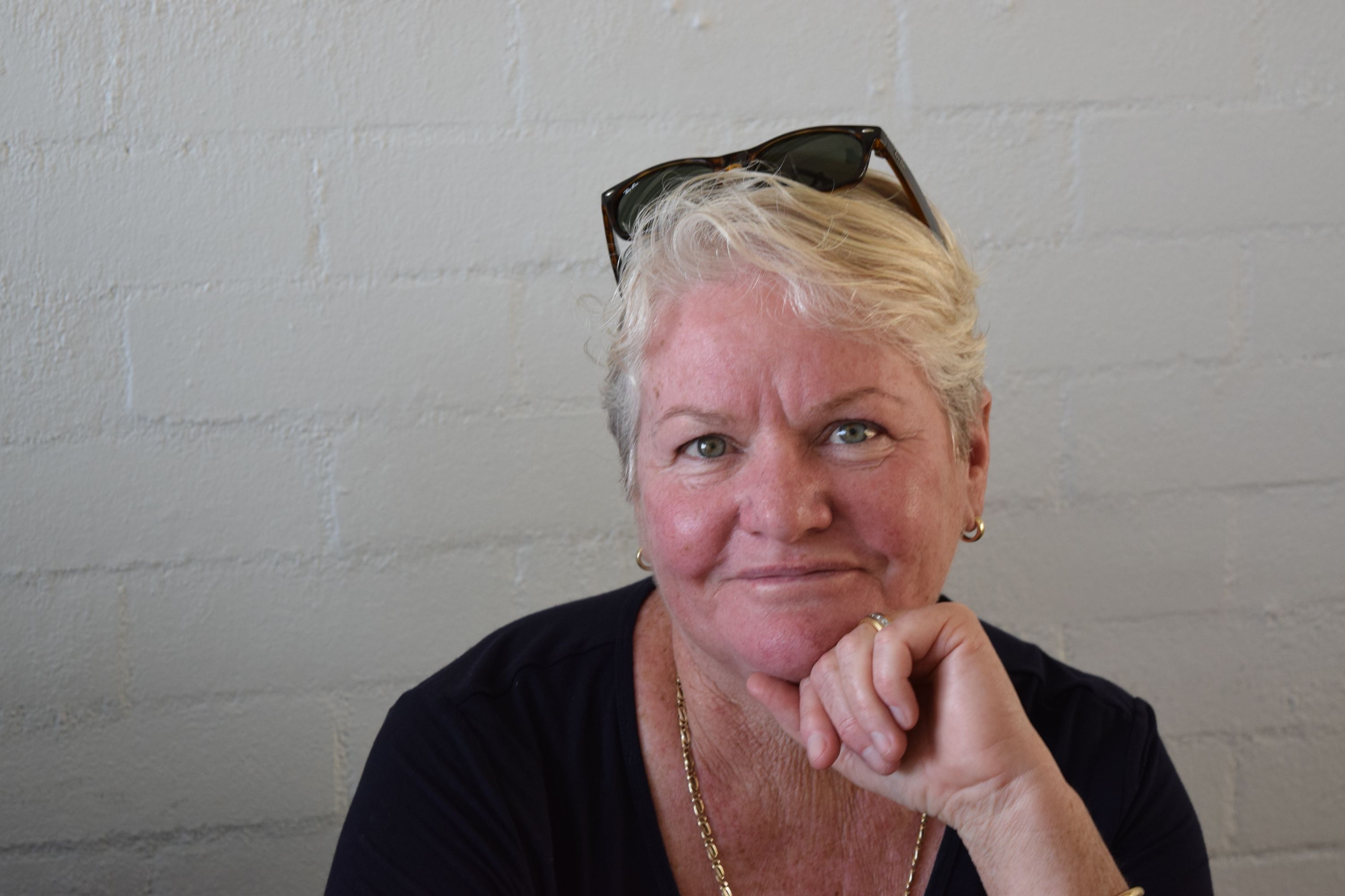 A middle-aged woman with short, blonde hair wears a dark shirt as she sits in front of a painted brick wall.