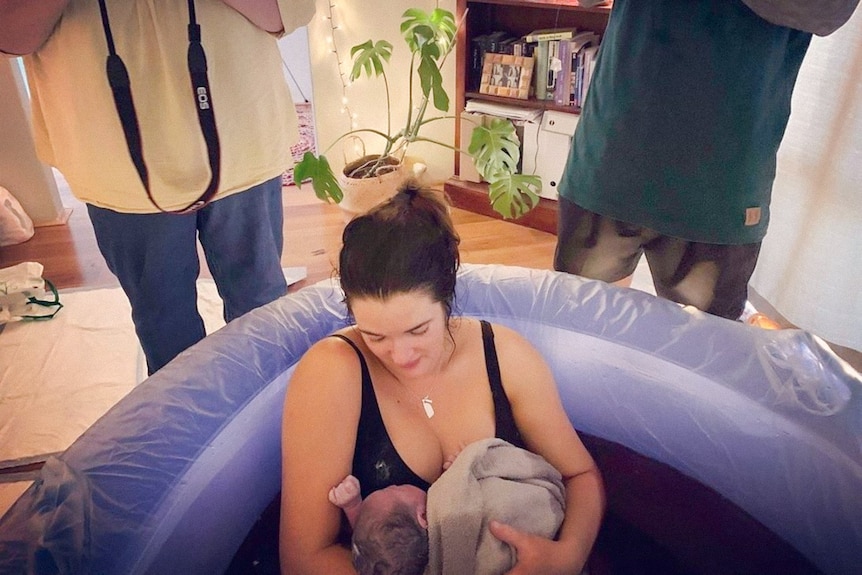 Laura holding her newborn baby while sitting in a birthing pool in a living room after her home birth.