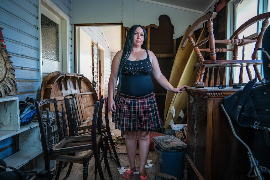 Woman wearing skirt and singlet stand among damaged furniture piled in a room