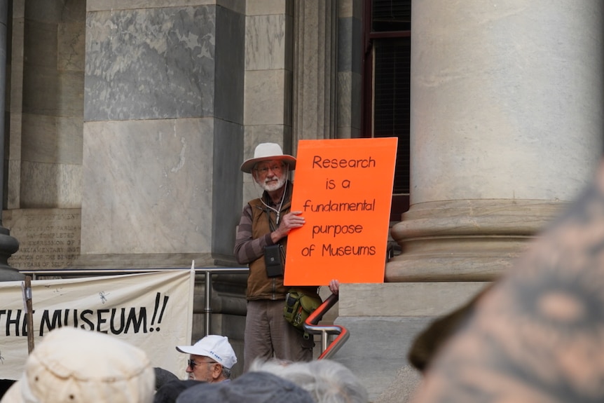 A protester holding up a sign that says "research is a fundamental purpose of museum".