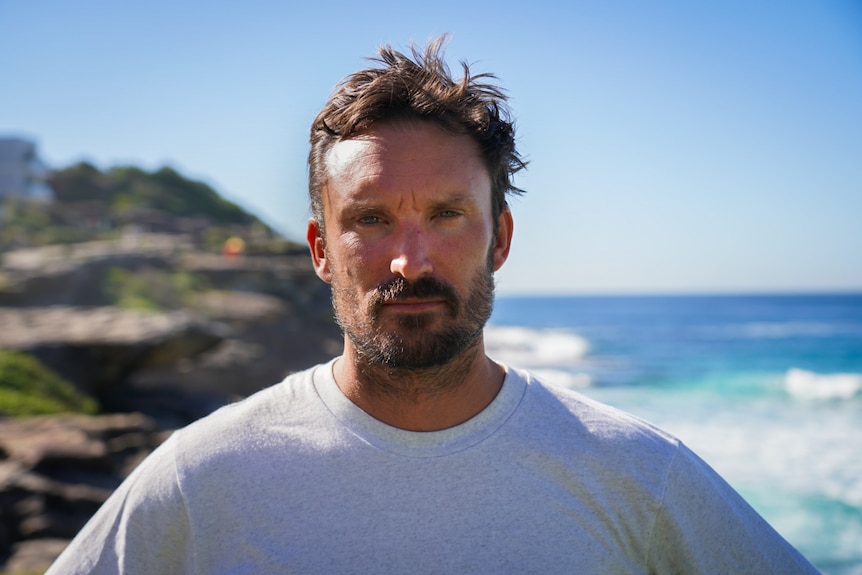 A man wearing a white shirt looks at the camera. In the background there is a beach and a cliff.
