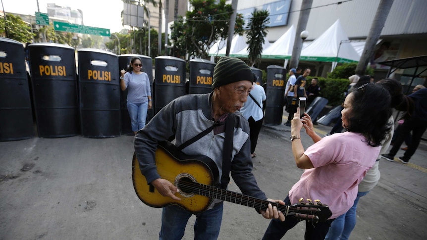 A man with a guitar plays as a woman behind him gets her portrait in front of a row of black riot shields.