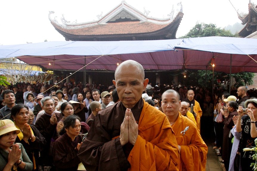 A monk walks with his hands together among dozens of followers at a temple