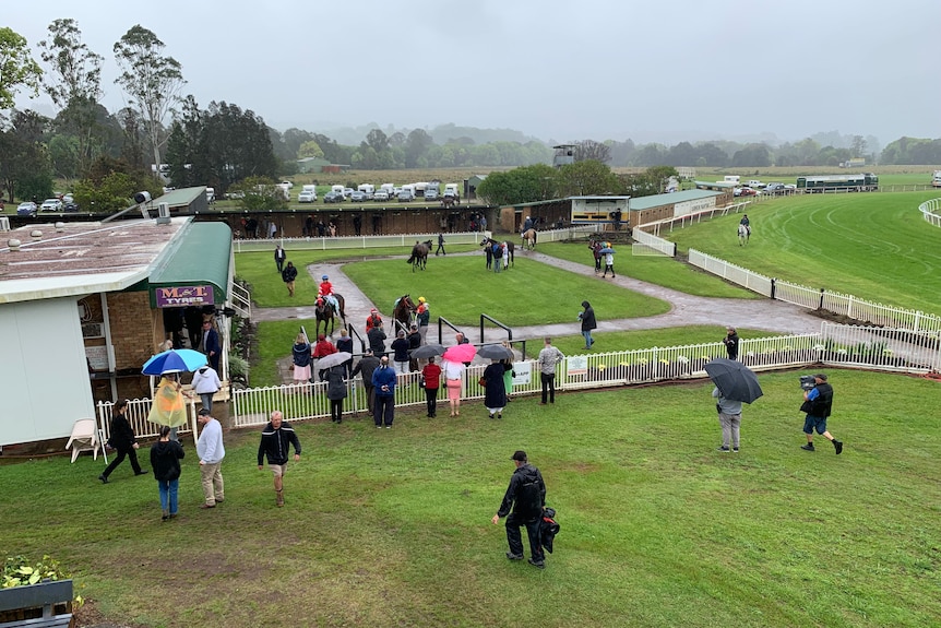 A high view of people at a mounting yard at a racing track. The grass is very green with patches of brown mud breaking through.