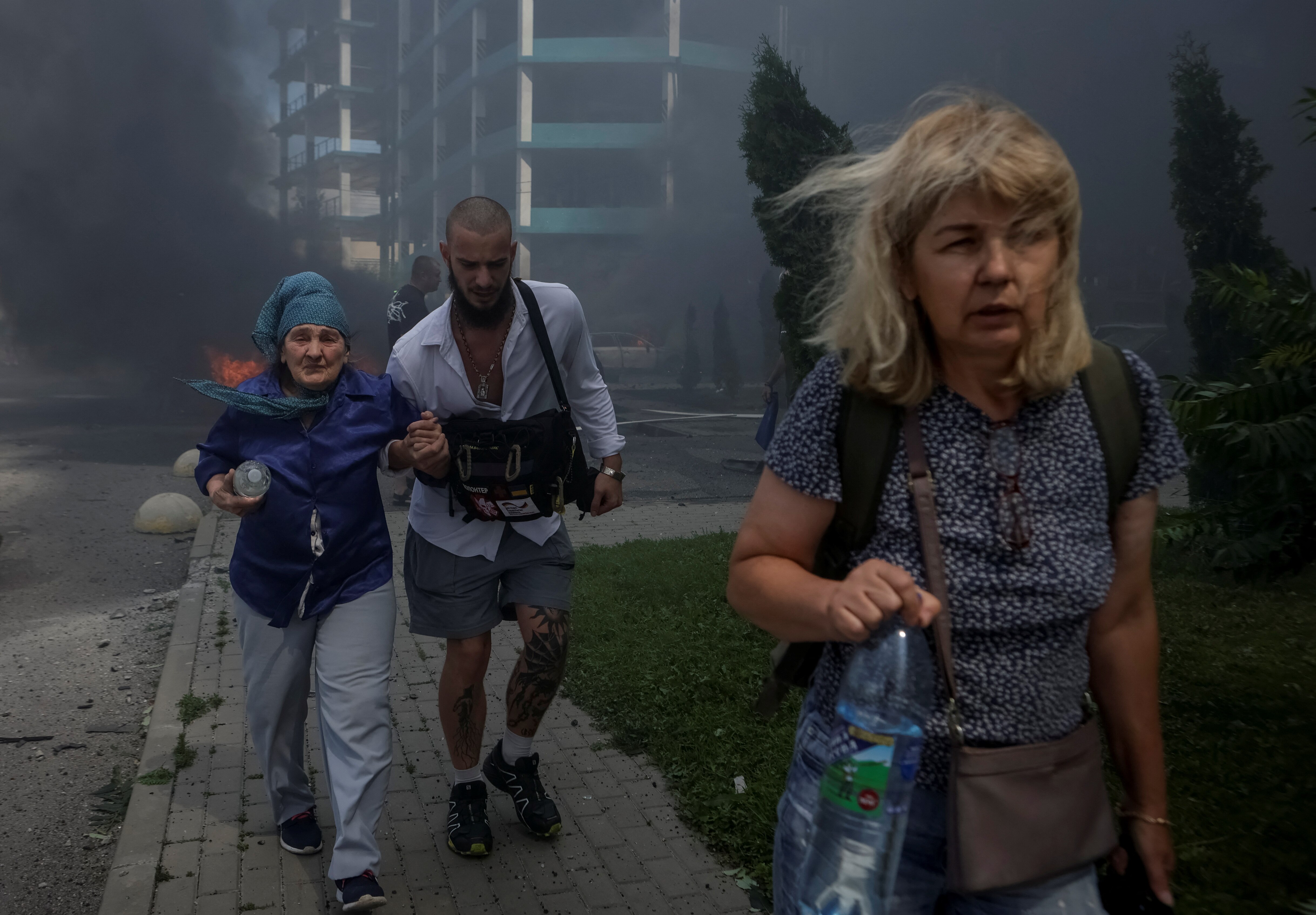 A middle aged woman and an elderly woman grasping the arm of a younger man walk away from smoke and flames behind them