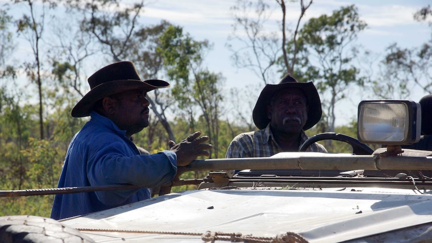 Two aboriginal men lean on a car chatting.