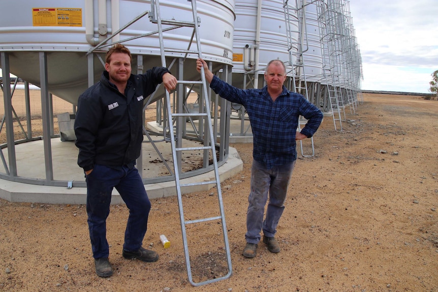 Jeff Munns and his son Dan stand along side a row of grain silos in a paddock.